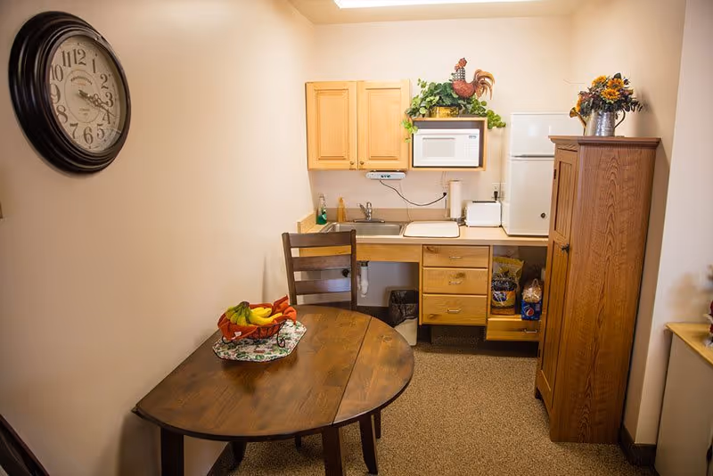 Small kitchenette and dining area with a round wooden table, wall clock, sink, microwave, mini fridge, and wooden cabinets.