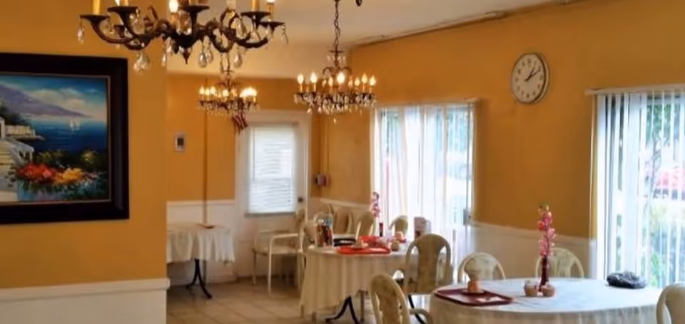 A dining room with round tables covered in white tablecloths, each table decorated with small flower vases. The room has yellow walls with white wainscoting, two chandeliers hanging from the ceiling, a wall clock, and large windows with vertical blinds letting in natural light. There is a framed painting on one wall and several chairs around the tables.