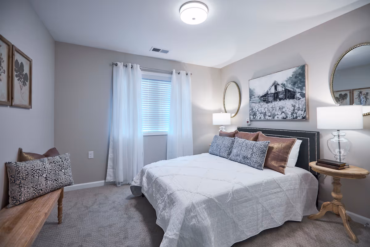 A cozy bedroom with a neatly made bed covered in a white quilt and decorated with multiple pillows. There is a wooden bench with a patterned cushion against one wall, two framed botanical prints above it, and a window with white curtains letting in natural light. On the right side of the bed, there is a round wooden side table with a glass lamp and two books. Above the bed hangs a black and white painting of a barn in a field, flanked by two round mirrors on the wall. The room has light gray walls and carpeted flooring.