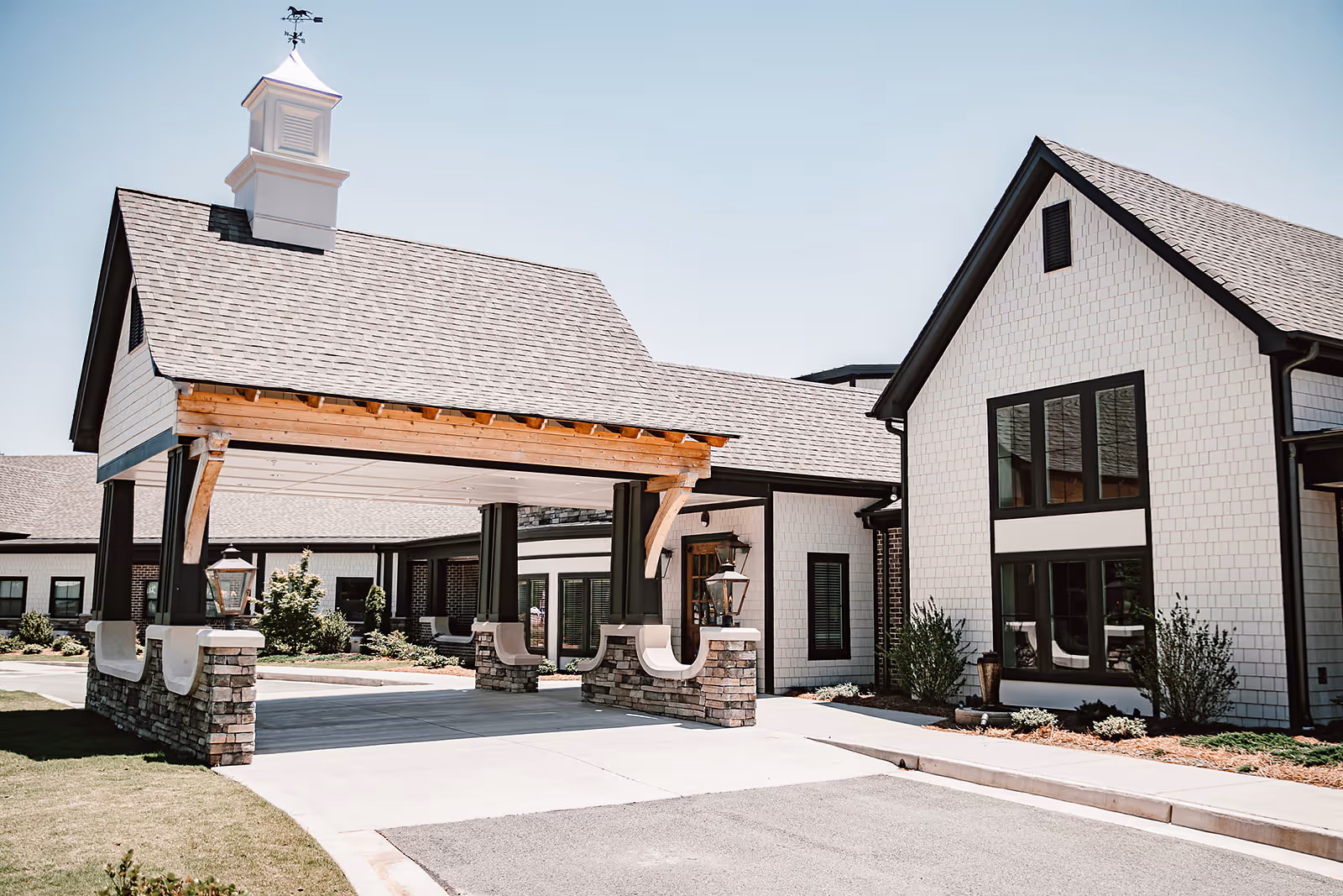 Exterior view of a senior living facility with a covered entrance supported by stone pillars and wooden beams. The building has white siding, large windows, and a cupola with a weather vane on the roof. Landscaping with shrubs and grass surrounds the entrance area.