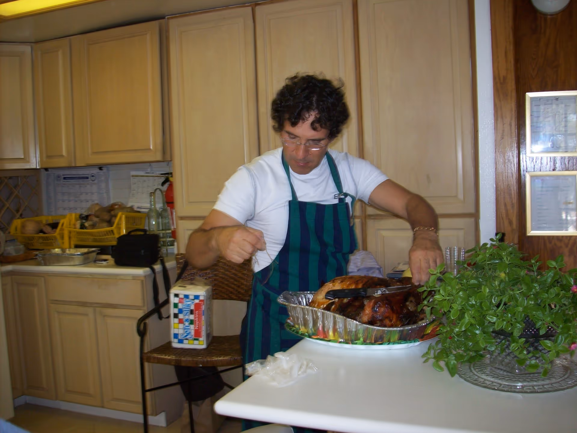 A man wearing a white shirt and a green and blue striped apron is carving a roasted turkey in a kitchen with light wood cabinets. There is a green leafy plant on the white countertop next to him, and kitchen items such as yellow baskets and a calendar are visible in the background.