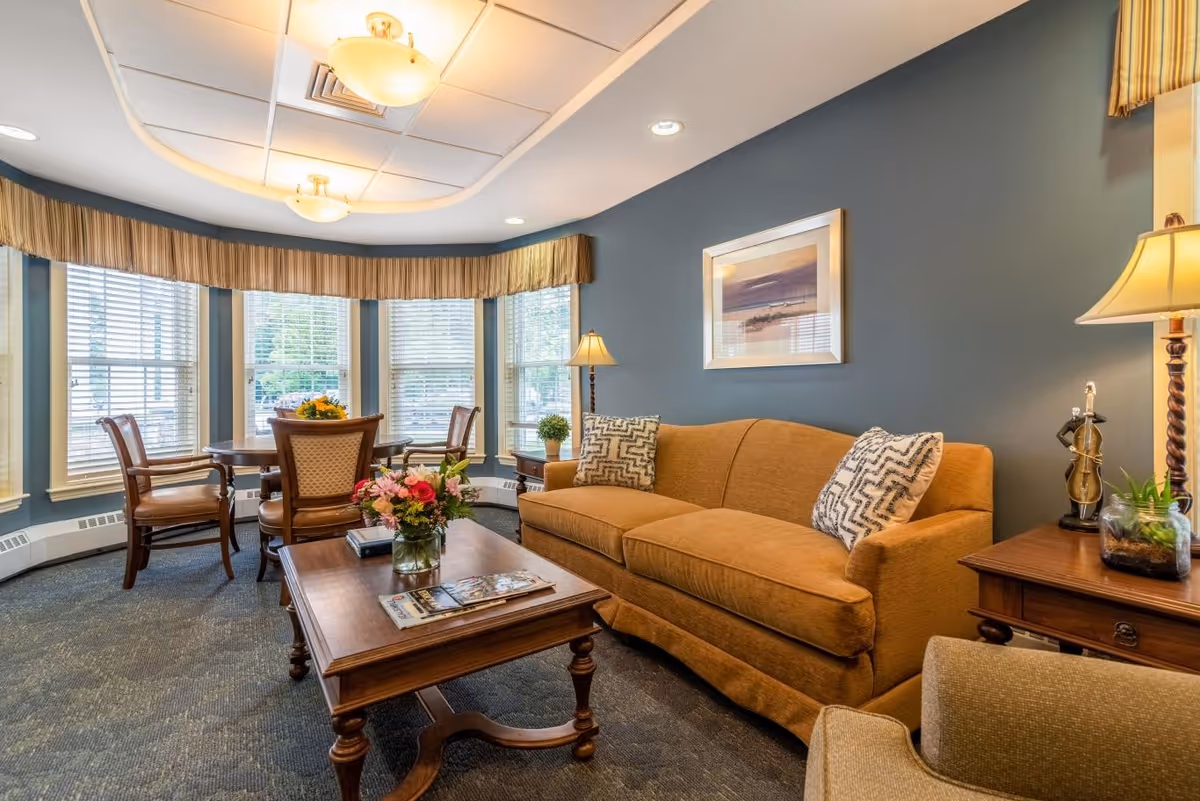 A cozy living room with a mustard yellow sofa adorned with patterned pillows, a wooden coffee table with a flower vase and magazines, a round dining table with four chairs near large windows with blinds, and warm lighting from ceiling fixtures and table lamps. The walls are painted blue and decorated with a framed picture.