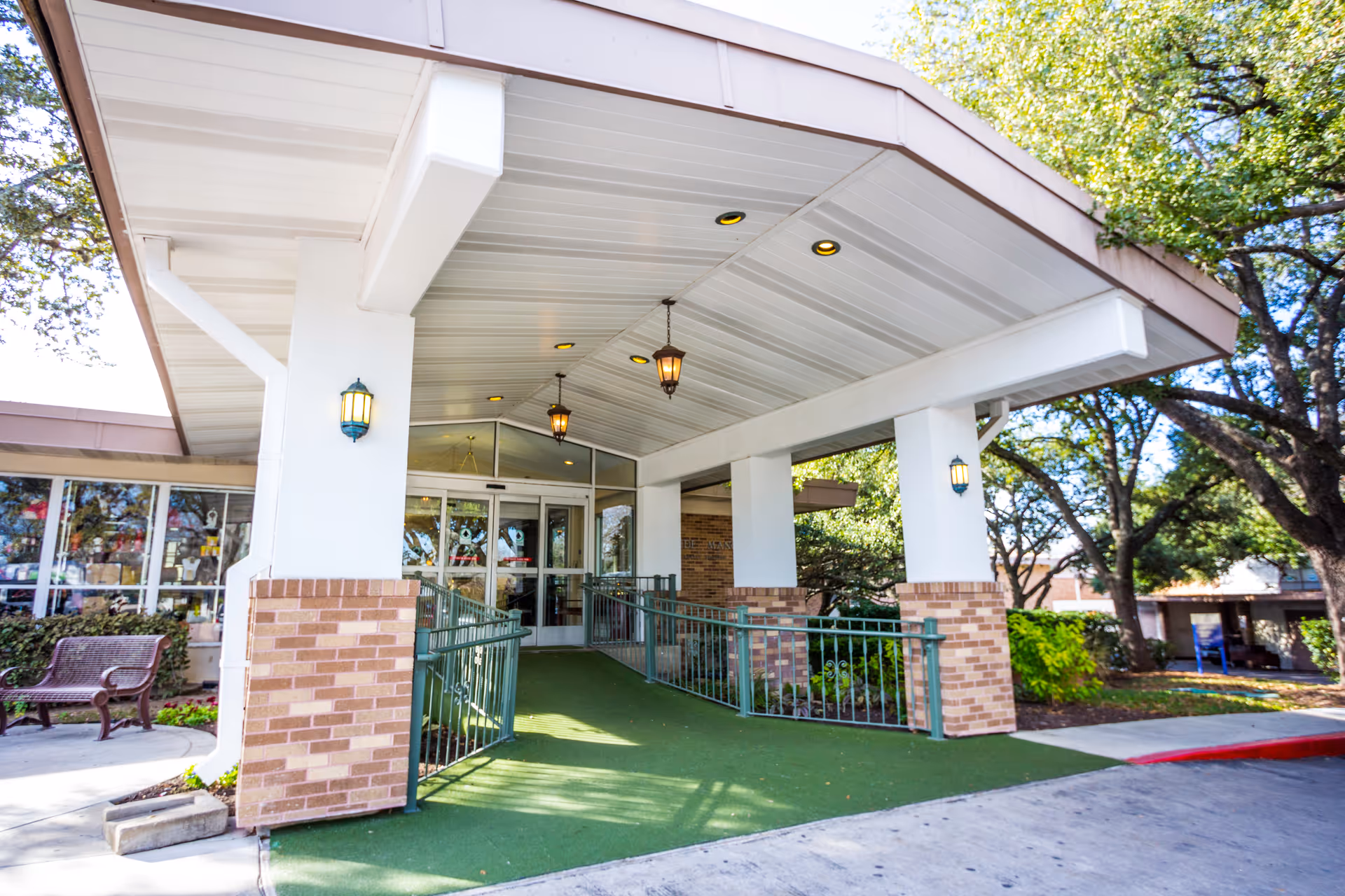 Entrance to a building with a covered walkway supported by white pillars with brick bases, green railings, and a green carpeted ramp leading to glass double doors. There are hanging lantern-style lights under the roof and trees surrounding the area.