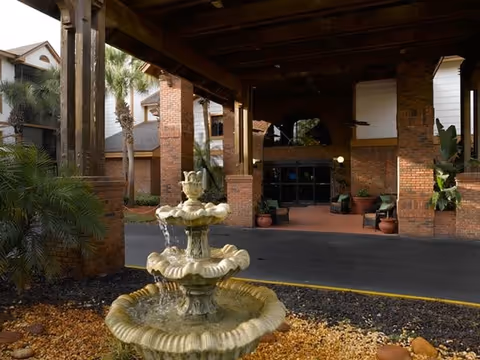 Outdoor view of the entrance area of a senior living facility with a multi-tiered water fountain in the foreground, surrounded by landscaping with rocks and plants. The building features brick pillars and a covered driveway with seating areas near the entrance.