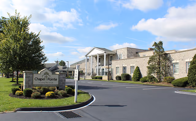 Exterior view of Chapel Pointe At Carlisle senior living community building with a stone facade, columns at the entrance, a driveway, and landscaped greenery including trees and bushes under a partly cloudy sky.
