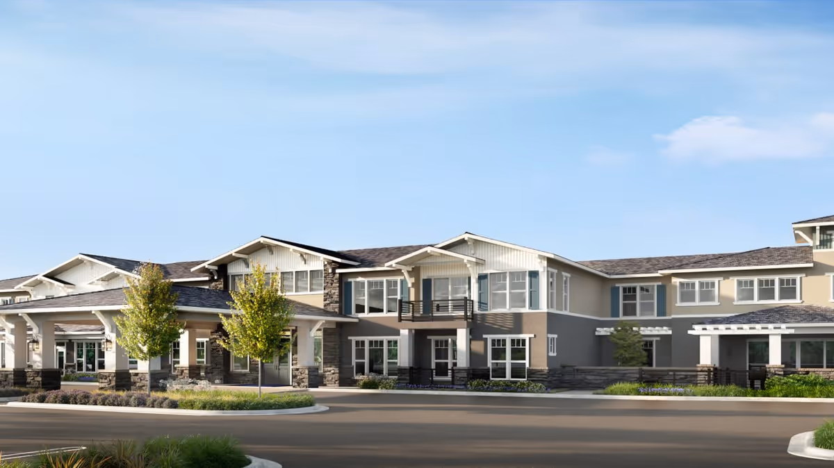 Exterior view of a modern senior living facility building with beige and gray walls, white trim, multiple windows, and a covered entrance. There are small trees and landscaped greenery in front of the building under a clear blue sky.