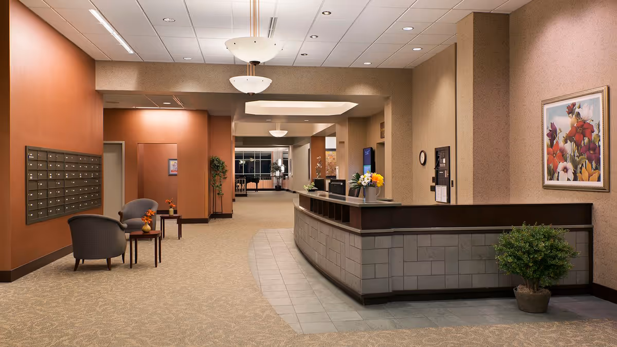 Interior view of a senior living facility lobby with a curved reception desk adorned with flowers, two gray chairs with small tables and flower vases, a wall of mailboxes, a potted plant, and framed floral artwork on the wall. The space is warmly lit with ceiling lights and has beige and brown tones.