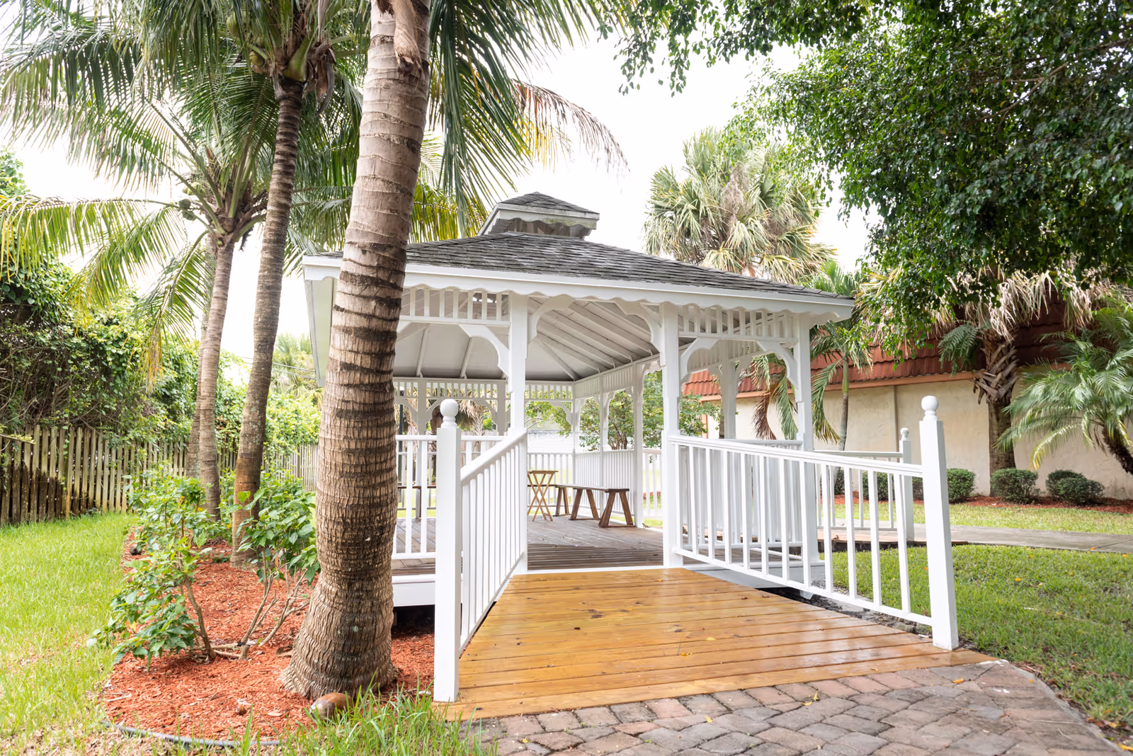 A white wooden gazebo with a ramp and seating surrounded by palm trees and landscaped grounds.