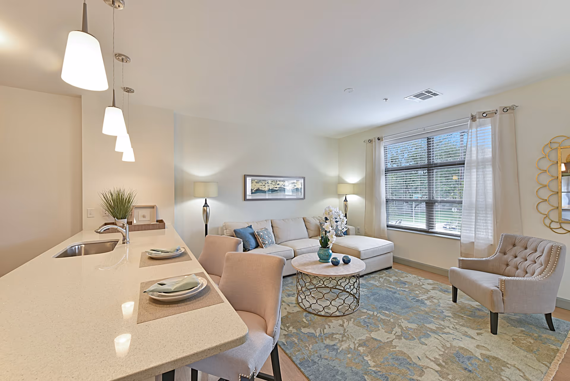 A bright and modern living room with a large window covered by sheer curtains, a beige sectional sofa with blue and patterned pillows, a round coffee table with decorative items, a beige armchair, and two floor lamps. Adjacent to the living area is a kitchen counter with a sink, two place settings, and three hanging pendant lights above.