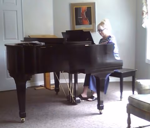 A woman with blonde hair and glasses is seated at a black grand piano in a carpeted room with white walls. There is a framed picture of a violin on the wall behind the piano, a closed white door to the left, and a window with curtains to the right. A cushioned chair is partially visible in the foreground on the right side.