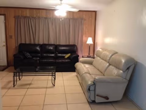 A living room with two sofas facing each other, one black leather and one beige leather recliner. There is a glass coffee table with a black metal frame between them. The room has tiled flooring, wood-paneled walls with a window covered by beige curtains, a ceiling fan with a light, and a floor lamp next to the beige sofa.