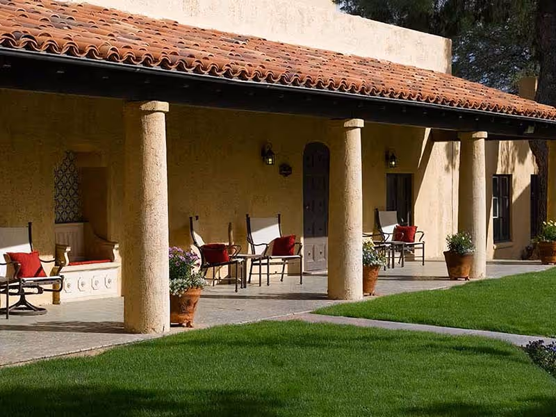 A shaded stucco porch with a tiled roof and columns overlooking a green lawn, furnished with chairs, potted plants, and red cushions.