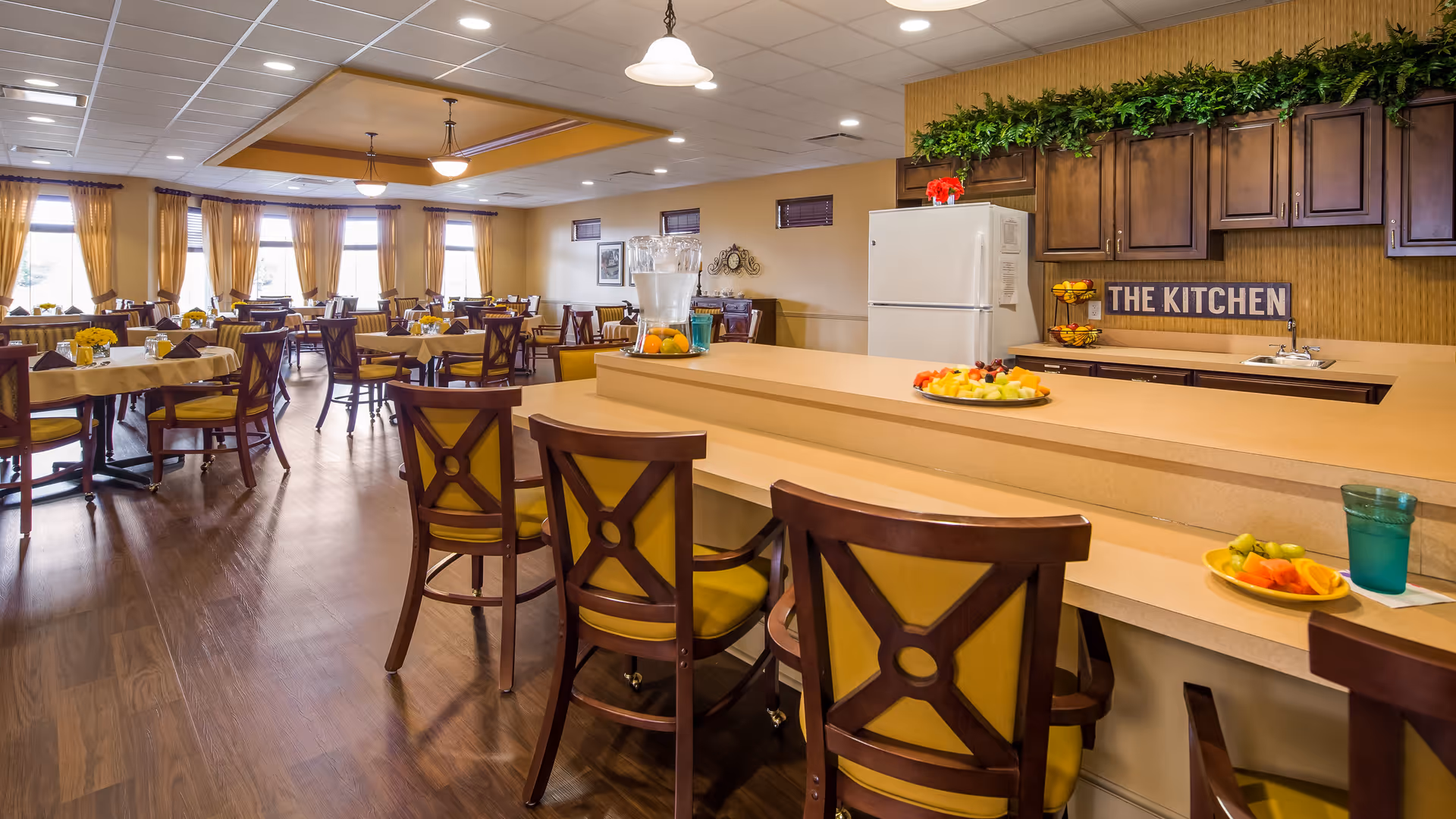 A spacious dining area with multiple tables and chairs arranged neatly. The room has large windows with yellow curtains allowing natural light to fill the space. In the foreground, there is a counter with bar stools, plates of fruit, and a water pitcher. Behind the counter, there is a kitchen area with wooden cabinets, a refrigerator, and a sign that reads 'THE KITCHEN'.