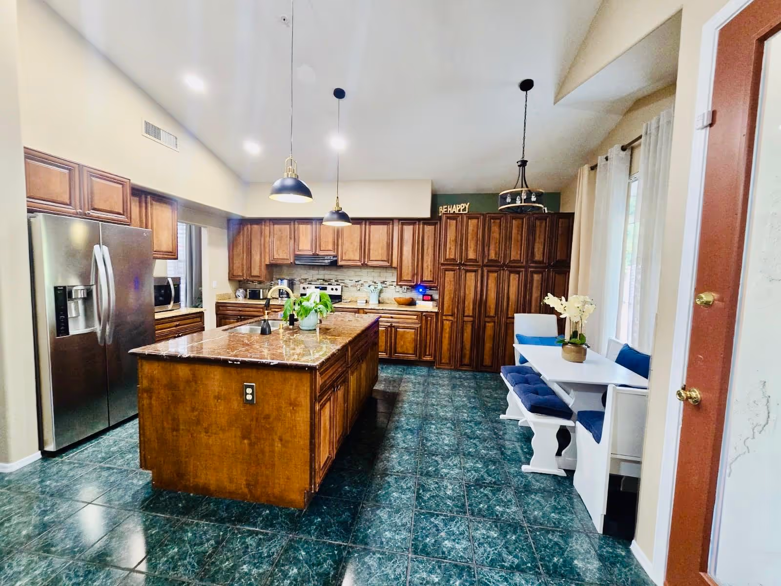 A spacious kitchen with dark green tiled floor, wooden cabinets, a large kitchen island with a marble countertop, and stainless steel refrigerator. There are two pendant lights hanging above the island. To the right, there is a small dining nook with a white table, blue cushioned bench, and a white chair. A door with a brass handle is visible on the far right.