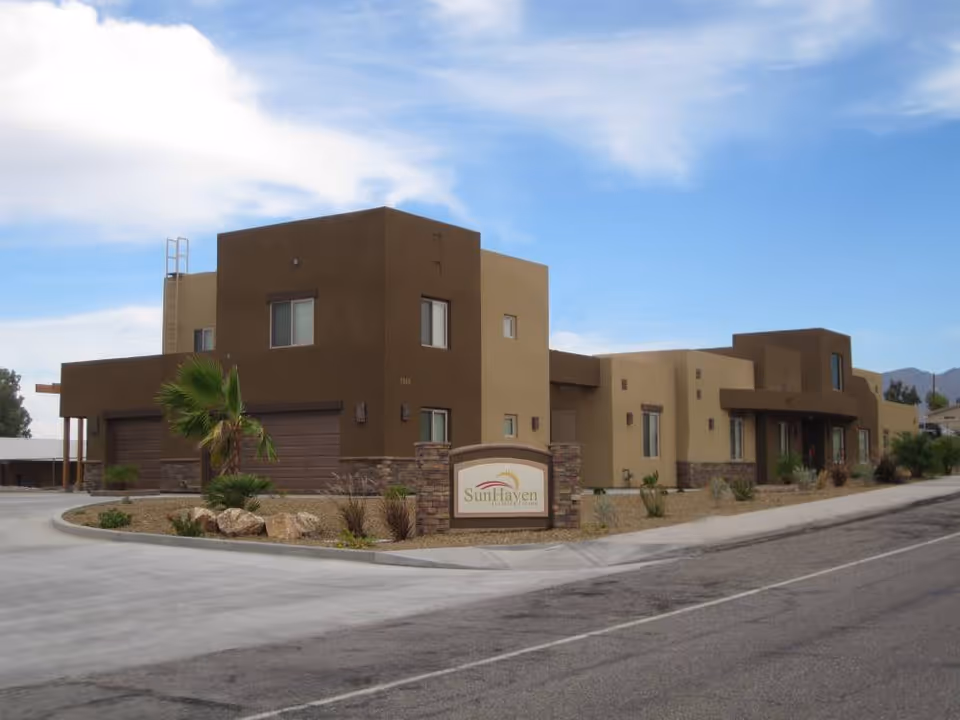 Exterior view of a modern, single-story assisted living facility with a flat roof and earth-toned walls. The building has several windows and a driveway leading to a garage. There is a sign in front that reads 'SunHaven Assisted Living' with some landscaping including small palm trees and rocks.