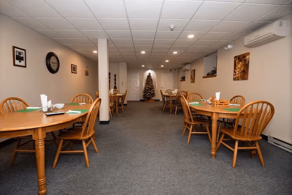 Interior view of a dining room with wooden tables and chairs arranged neatly. The room has a carpeted floor, beige walls with framed pictures, and a Christmas tree decorated at the far end near two white doors. The ceiling has recessed lighting and there is an air conditioning unit mounted on the wall.
