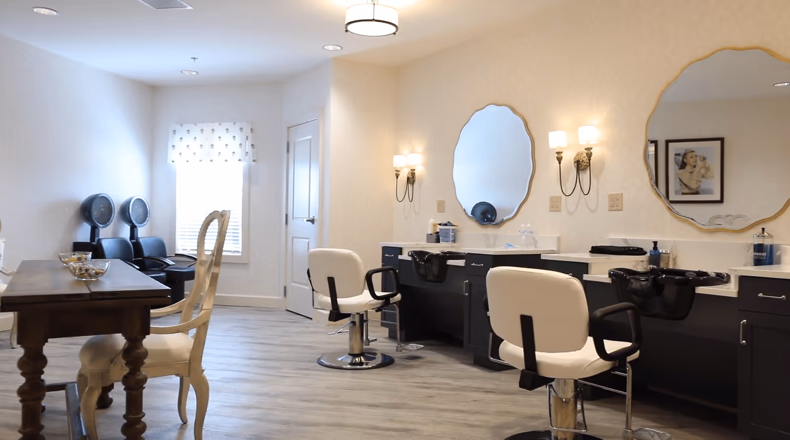 A bright and clean salon room with two white salon chairs in front of black counters with sinks and round mirrors on the wall. There are wall sconces with lights above the counters. To the left, there is a wooden table with two white chairs and two black hair dryer chairs near a window with a white curtain.