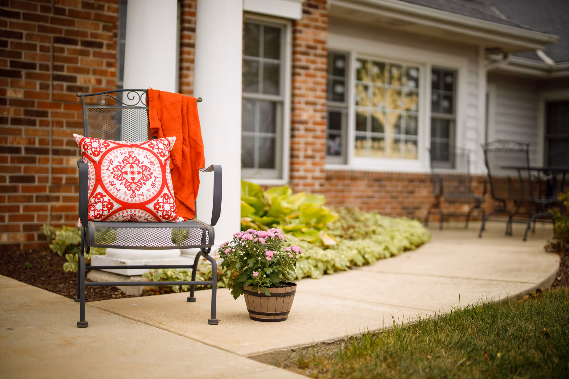 Outdoor patio area with a metal chair featuring a red and white patterned cushion and a red blanket draped over the back. A potted plant with pink flowers is placed on the concrete pathway next to the chair. The background shows a brick and siding exterior of a building with windows and additional metal chairs and a table.