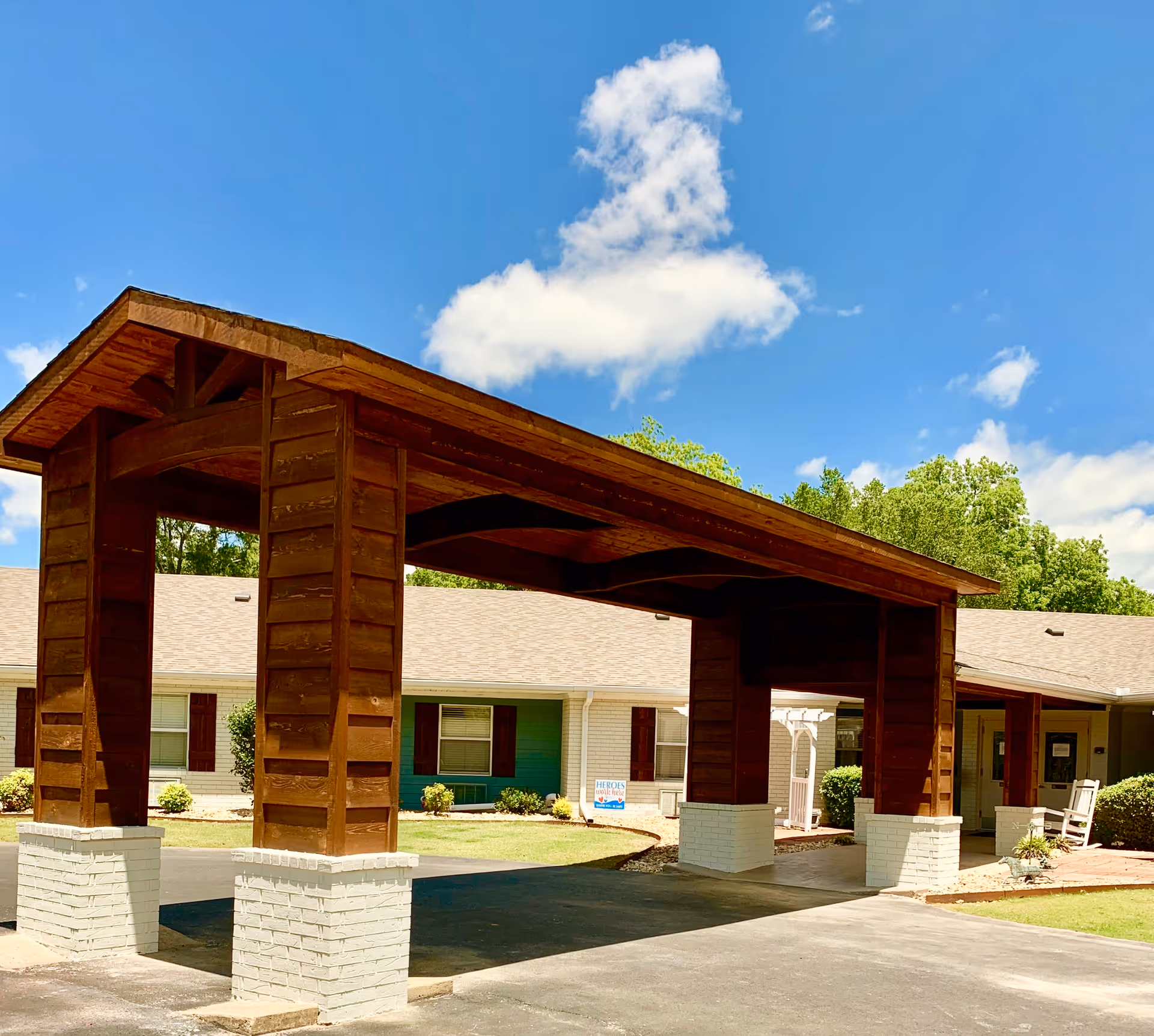 Entrance area of Village Park of Conway, Inc. featuring a large wooden covered driveway structure with white brick bases, a single-story building with beige siding and brown shutters, green bushes, and a bright blue sky with scattered clouds.