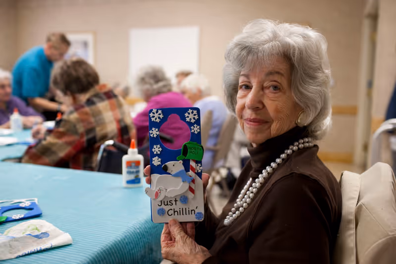 An elderly woman with gray hair and a pearl necklace is sitting at a table in a senior living facility craft room, holding up a decorated door hanger featuring a polar bear, snowflakes, and the text 'Just Chillin''. Other seniors are engaged in crafts in the background.