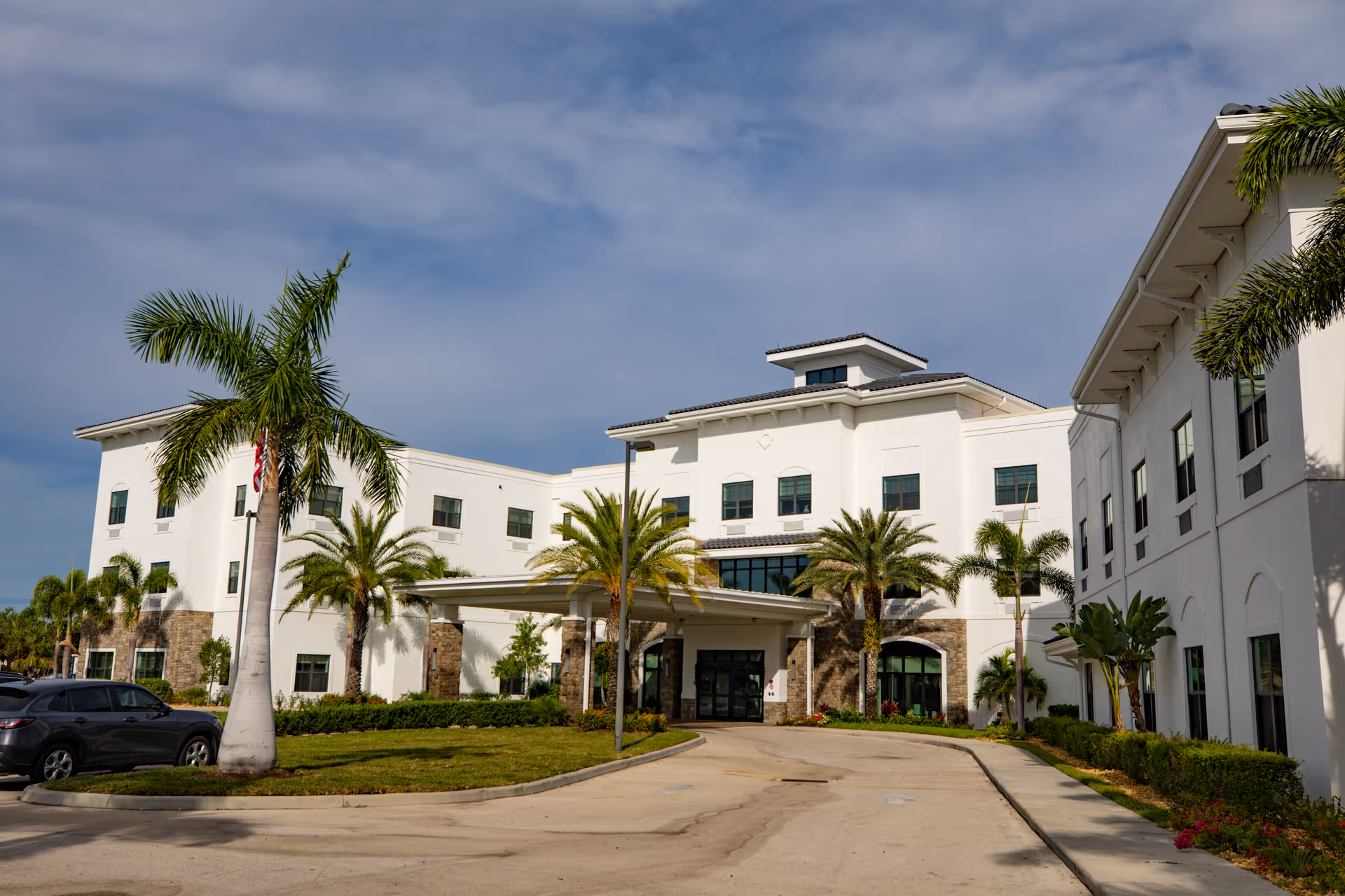 Exterior view of a white multi-story senior living facility building with palm trees and a driveway leading to the entrance under a partly cloudy sky.