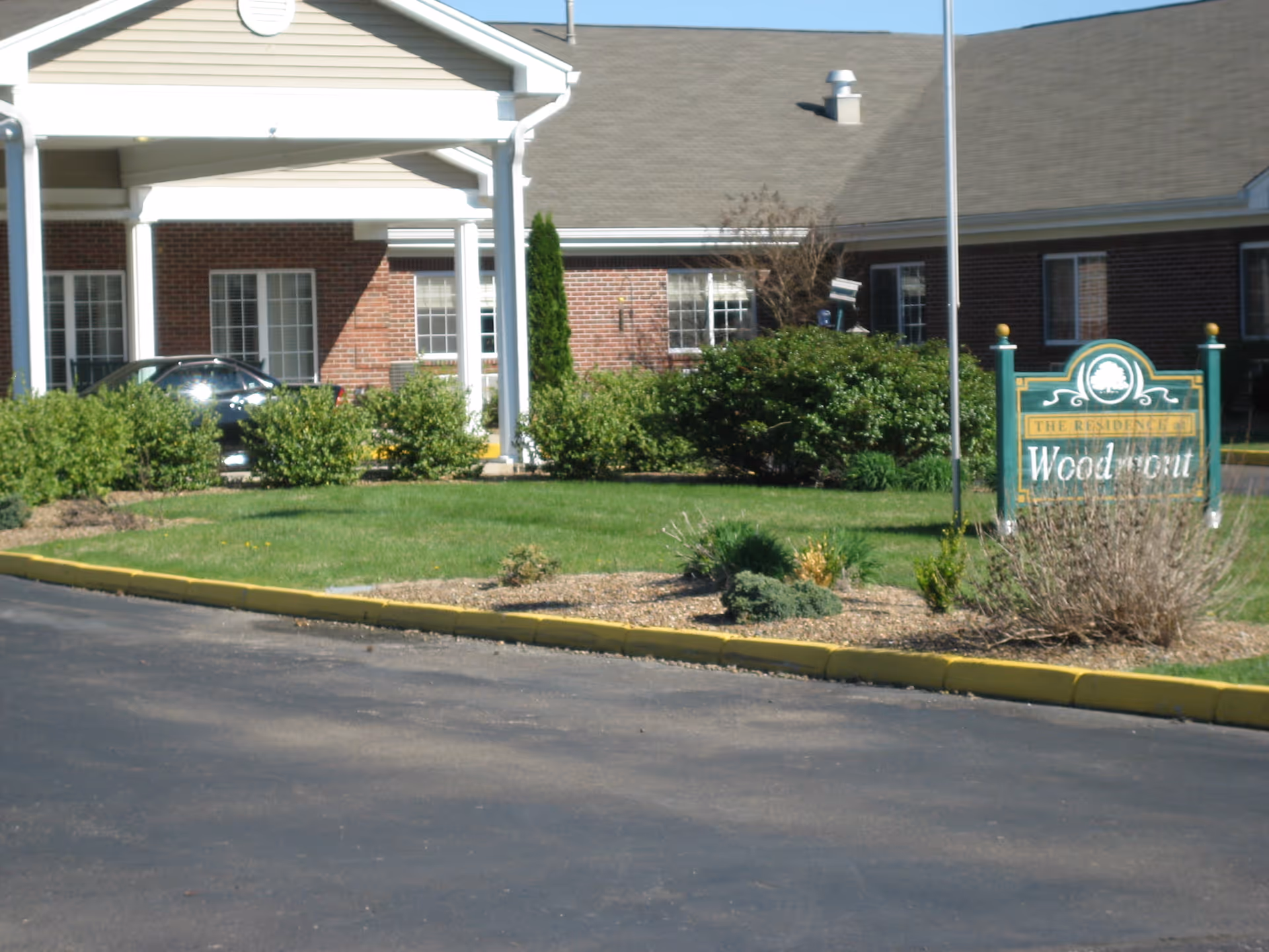 Front entrance of a brick senior living building with a covered portico, landscaped lawn, and a green Woodmont sign.