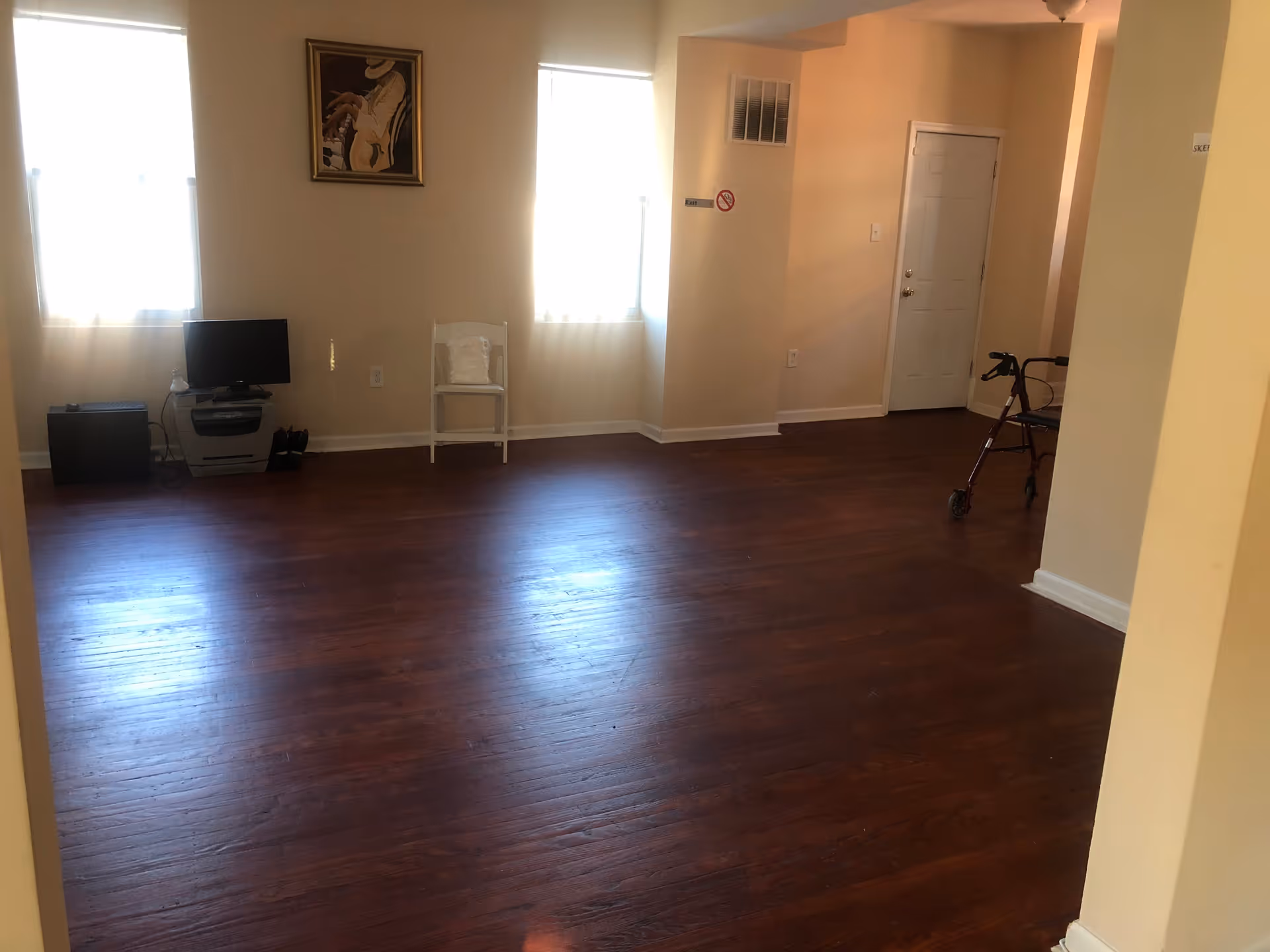 Spacious interior room with hardwood floors, two windows, a small TV and chair against the far wall, and a walker near a door.