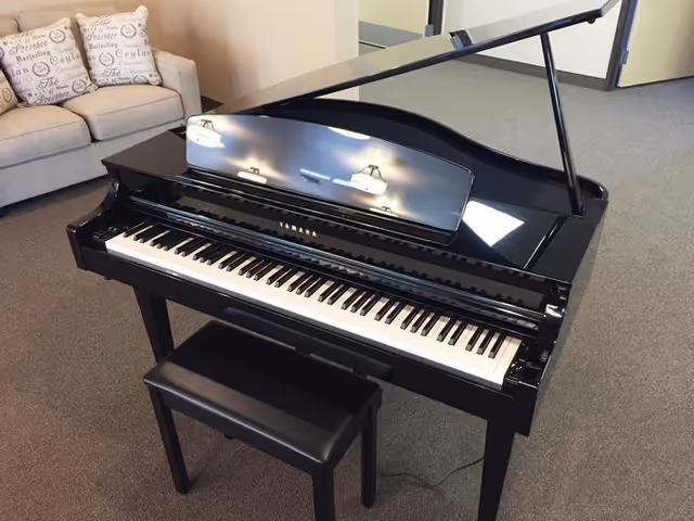 A black Yamaha grand piano with its lid open, accompanied by a black piano bench, placed on a carpeted floor in a room with a beige sofa and decorative pillows in the background.