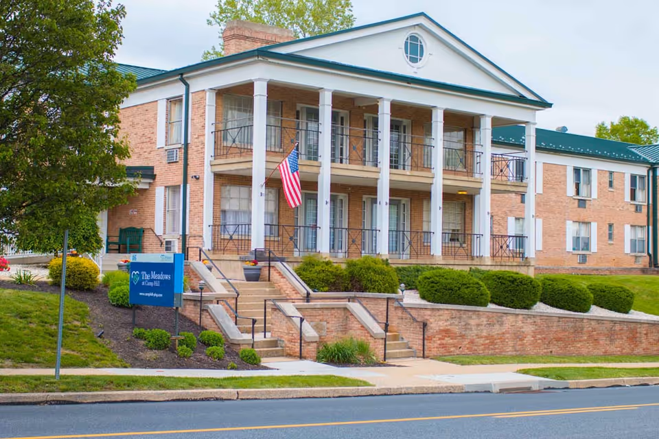 Two-story brick senior living building with white columns and balconies, front steps, landscaped grounds and an American flag.