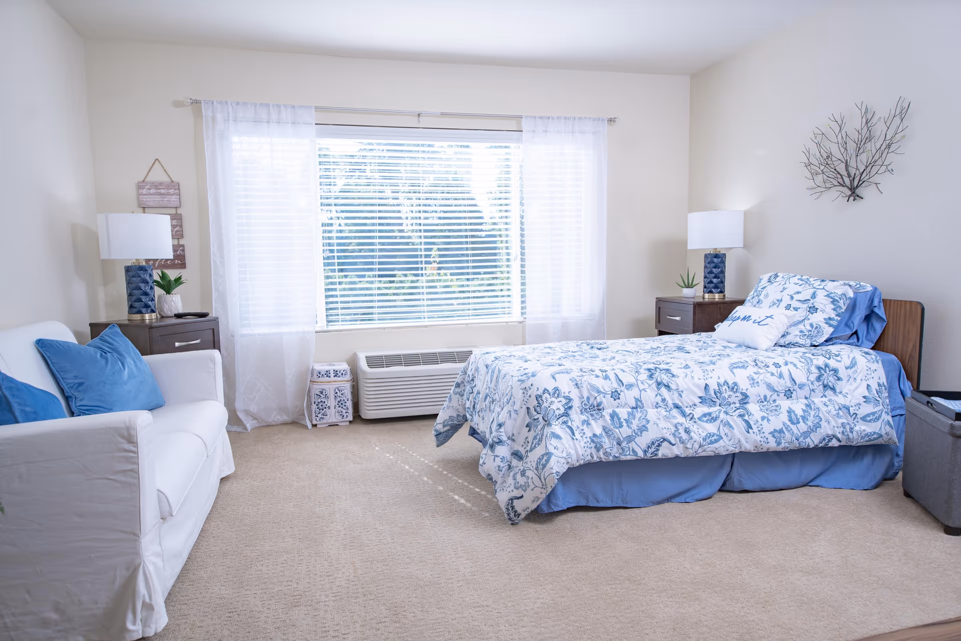 Bright, airy bedroom with a single bed in blue floral bedding, a white sofa, nightstands, and a large window with blinds.