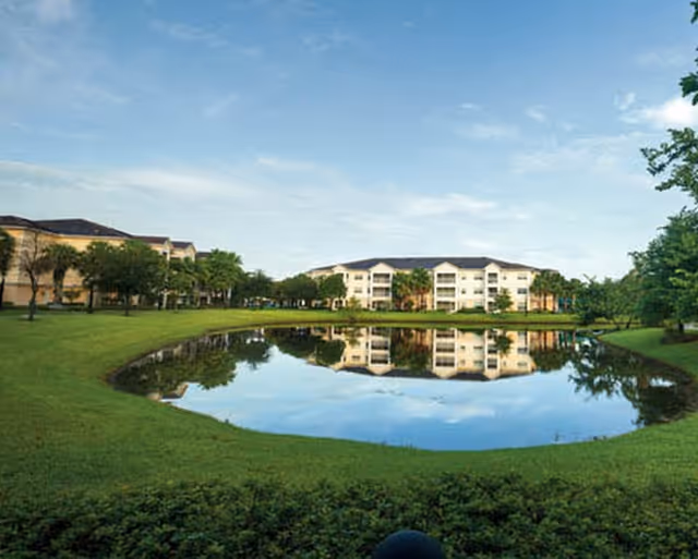 Multi-story senior living buildings reflected in a pond with surrounding lawns and trees under a blue sky.