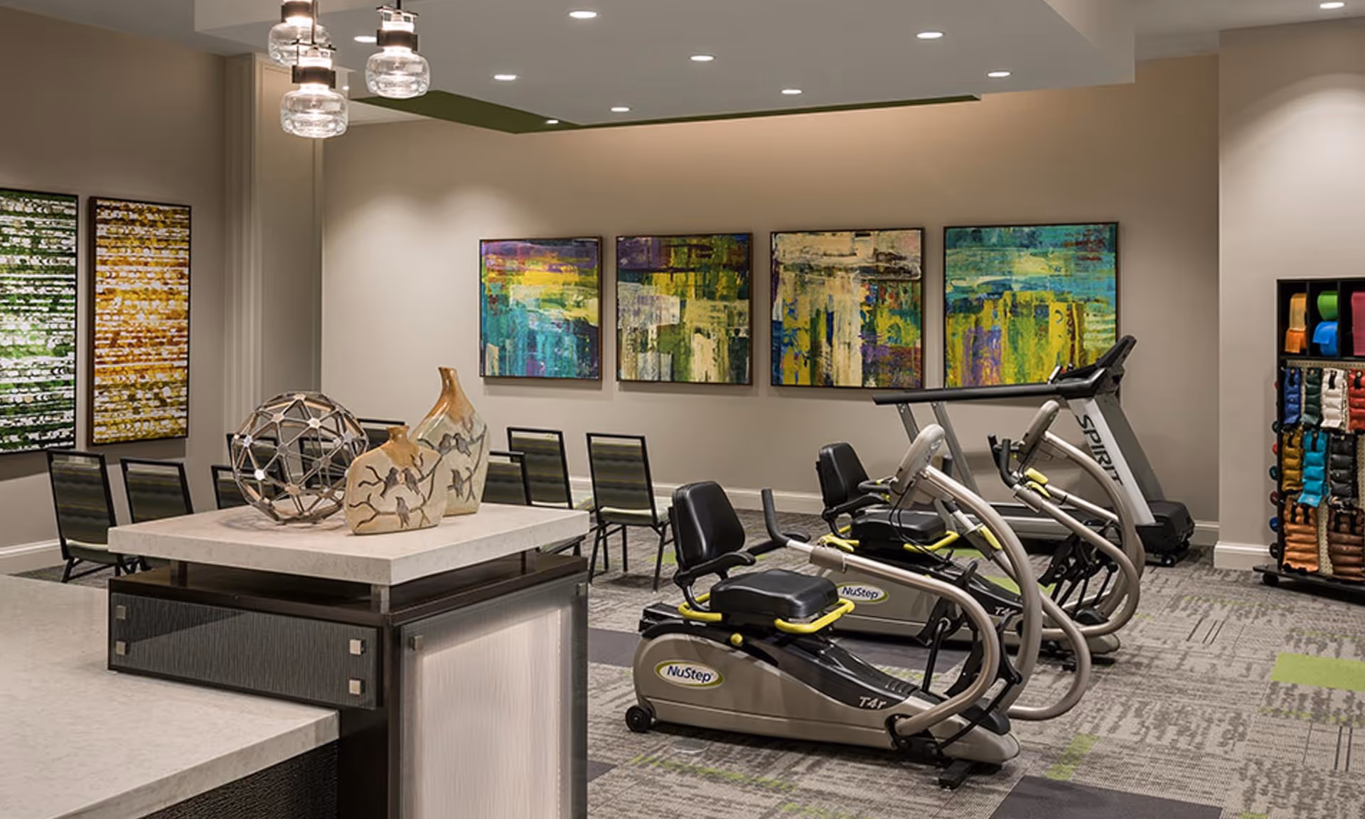 Well-lit interior fitness room with several recumbent cardio machines and a treadmill, seating area, decorative vases on a counter, and colorful abstract wall art.