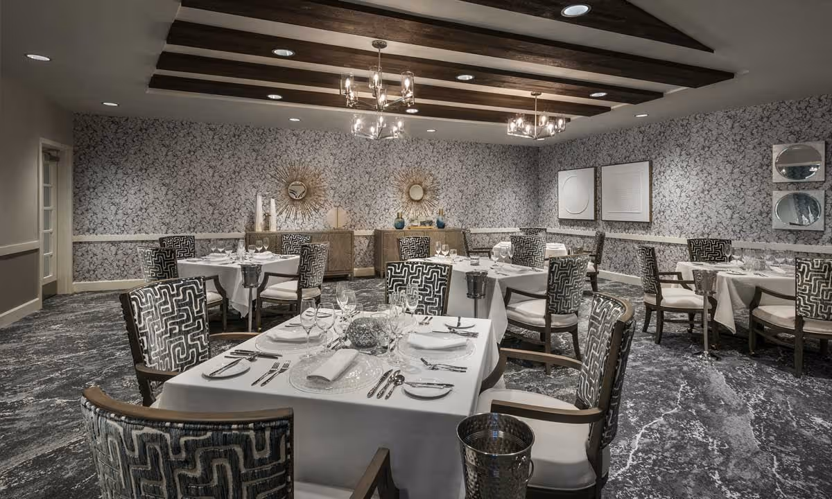 Elegant dining room with multiple tables set with white tablecloths, glassware, and silverware. The room features patterned chairs, decorative mirrors and artwork on the walls, and wooden ceiling beams with modern chandeliers providing lighting.