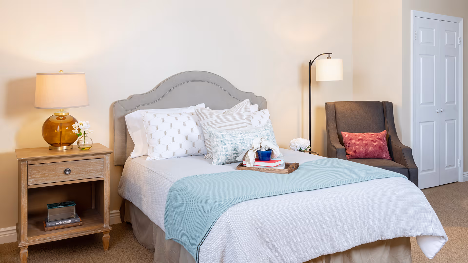 A neatly made bed with white and patterned pillows and a light blue throw blanket in a bedroom. Next to the bed is a wooden nightstand with a lamp and a small flower vase. There is a dark upholstered armchair with a red cushion and a floor lamp beside it. A white closet door is visible in the background.