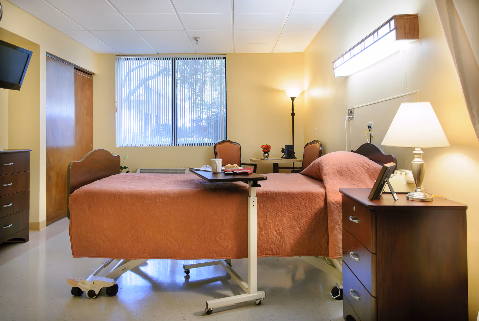 A well-lit senior care facility bedroom with a hospital-style bed covered in a brown quilt. The room has a window with vertical blinds, a wooden dresser with a lamp, a small table with two chairs, and a floor lamp in the corner. A TV is mounted on the wall, and there is a sliding wooden door.
