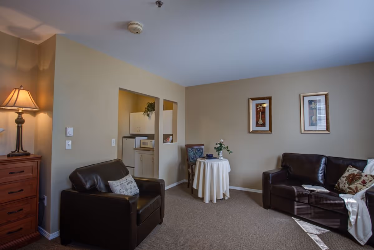 A cozy living room area with a dark brown leather armchair and sofa, a small round table covered with a white tablecloth, and a wooden chest of drawers with a lamp on top. The walls are beige with two framed pictures hanging. A small kitchen area with a refrigerator and microwave is visible through two open wall cutouts.