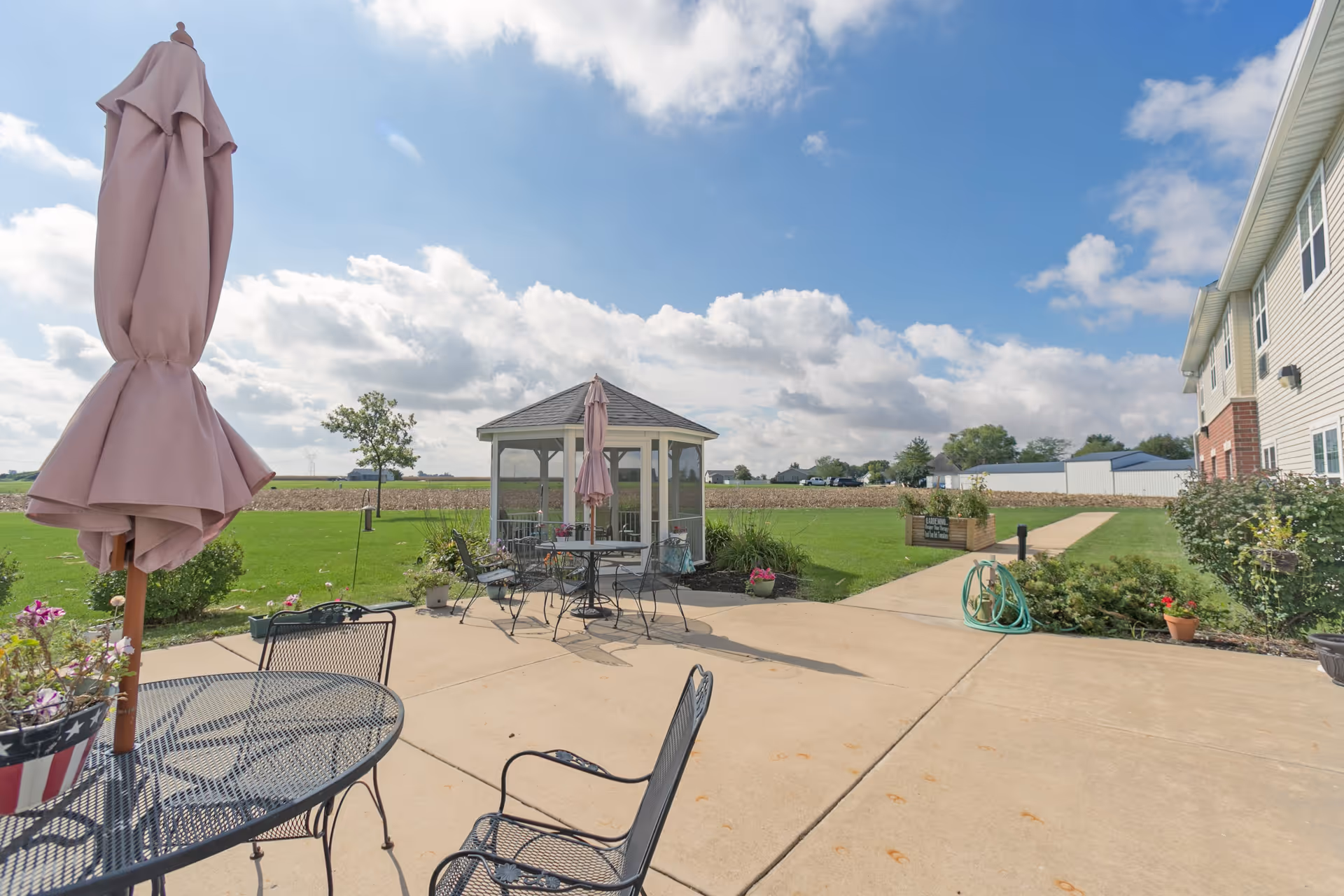 Outdoor patio area at Heritage Woods of Dwight with metal tables and chairs, closed pink umbrellas, a small gazebo, green lawn, and a clear blue sky with some clouds.