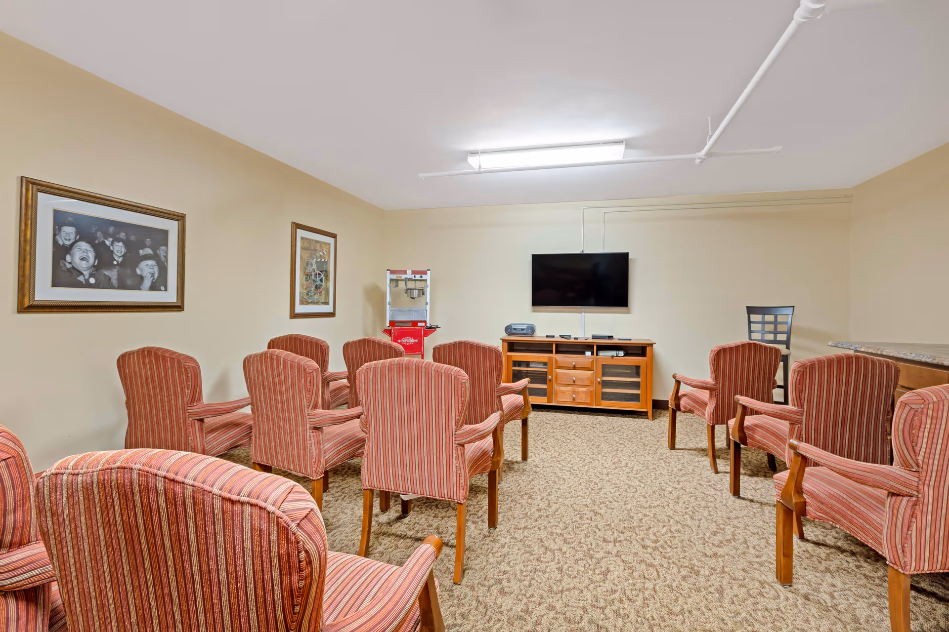 A small common room with several red striped armchairs arranged in rows facing a wall-mounted flat screen TV. Below the TV is a wooden cabinet with drawers and shelves holding electronic devices. On the left wall, there are two framed pictures, and a popcorn machine is positioned in the corner. The room has beige walls and carpeted flooring.