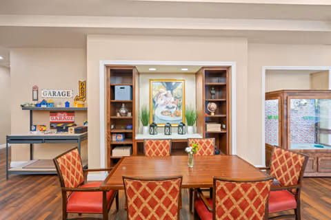 A dining area with a wooden table surrounded by six red and gold patterned chairs. Behind the table is a built-in shelving unit with decorative items and a framed painting of people dining. To the left, there is a small cart with vintage-style garage signs and decor. The room has wooden flooring and beige walls.