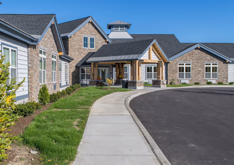 Exterior view of a single-story building with stone and siding facade, a covered entrance with wooden beams, a curved driveway, and a sidewalk surrounded by green grass and small shrubs under a clear blue sky.