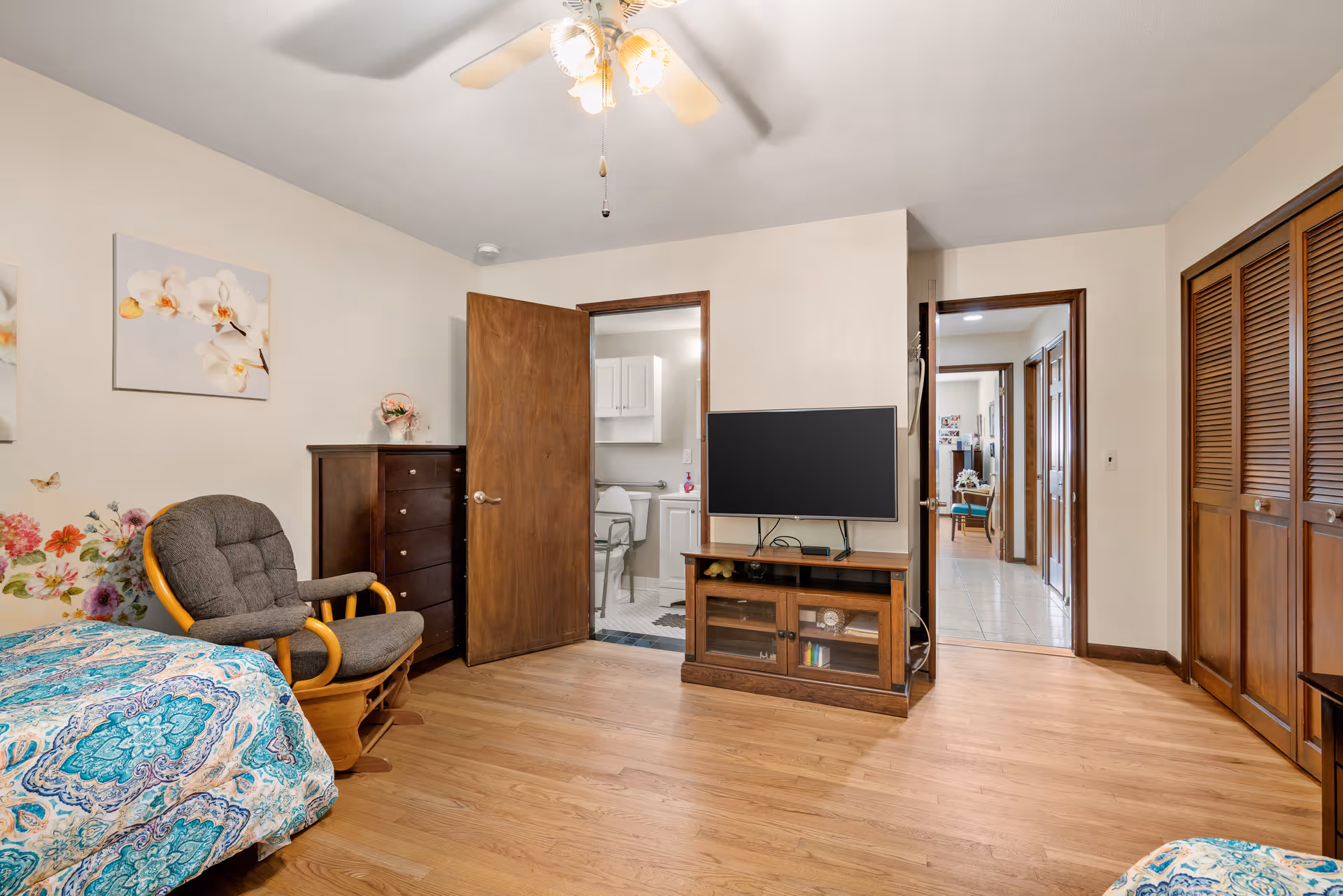 A cozy bedroom with wooden flooring, a ceiling fan with lights, a patterned bedspread, a cushioned rocking chair, a tall dark wooden dresser, a flat-screen TV on a wooden stand, and an open door leading to a bathroom with white cabinets and a toilet. Another open door leads to a hallway with tile flooring and additional rooms visible.