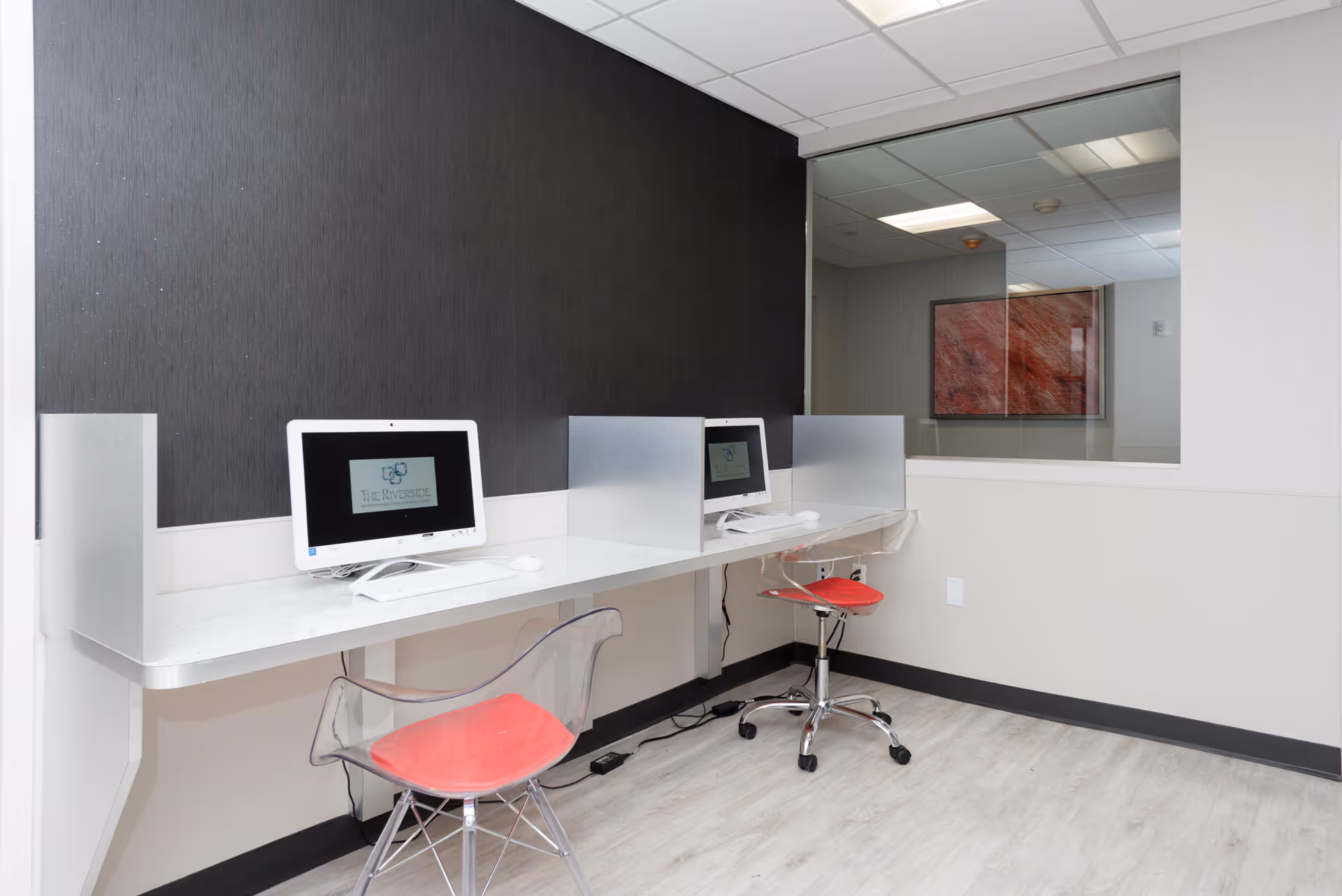 A small computer workstation area with two white desktop computers on a white counter. Each computer station is separated by a frosted glass divider. There are two transparent chairs with red cushions in front of the computers. The room has light-colored walls, a black accent wall behind the computers, and light wood flooring. A large window on the right side shows a view into another room with a framed artwork on the wall.