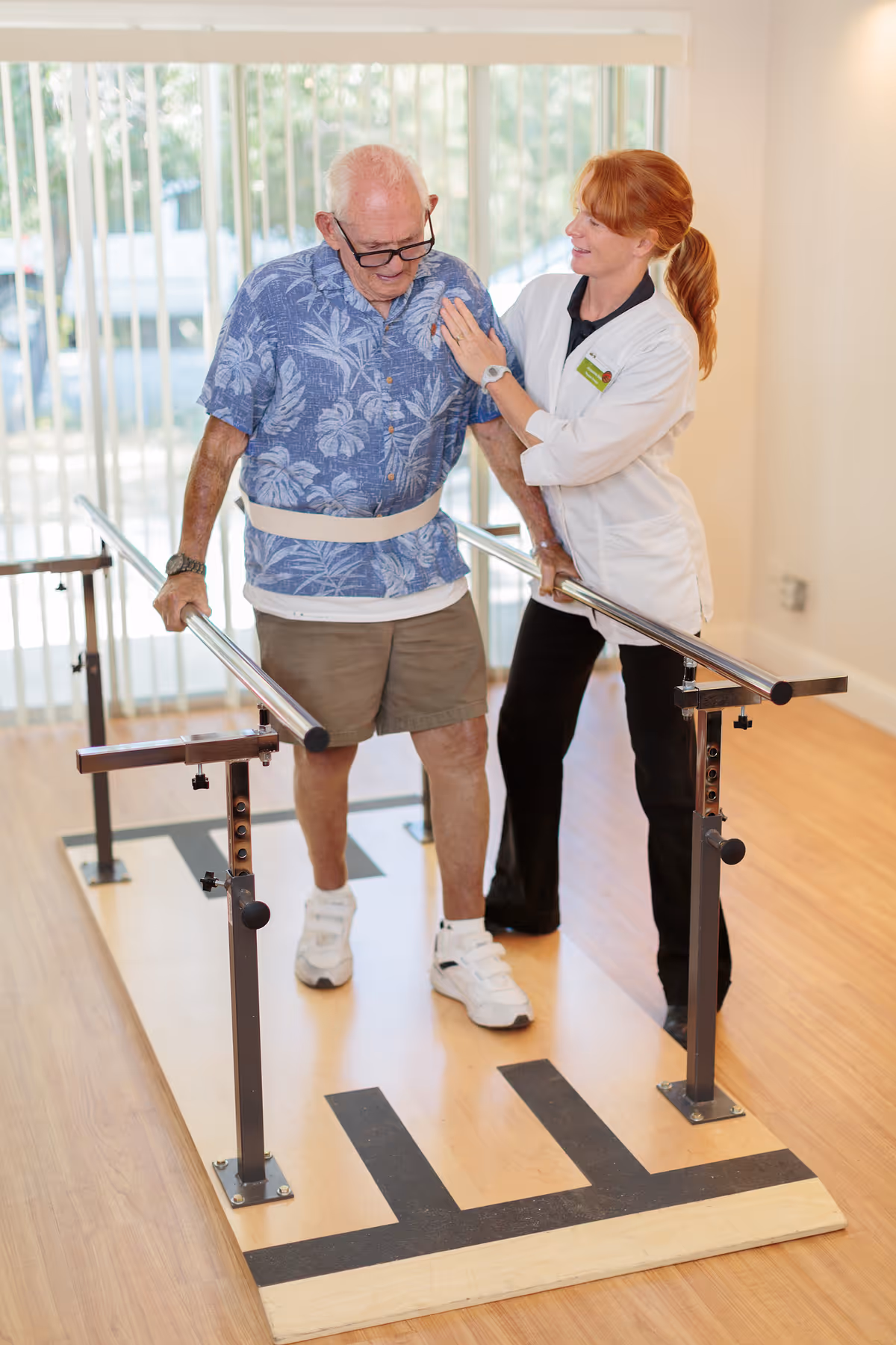 An elderly man wearing a blue floral shirt and beige shorts is walking between parallel bars for physical therapy, assisted by a female healthcare professional in a white coat who is supporting him by the shoulder. The room has wooden flooring and large vertical blinds covering the windows in the background.