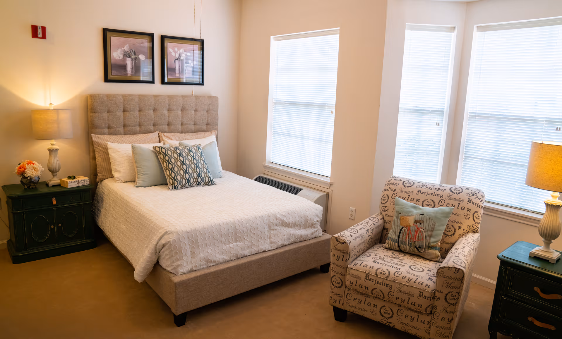 A cozy bedroom with a beige upholstered bed featuring white and patterned pillows. There are two framed floral pictures above the bed. On either side of the bed are dark green nightstands with lamps and decorative items. Next to the bed is a patterned armchair with a decorative pillow, positioned near three large windows with blinds.