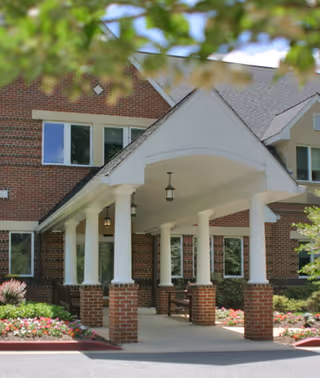 Entrance of a brick building with white columns supporting a covered porch. There are flower beds with colorful flowers on either side of the walkway leading to the entrance. Trees with green leaves partially frame the top of the image.