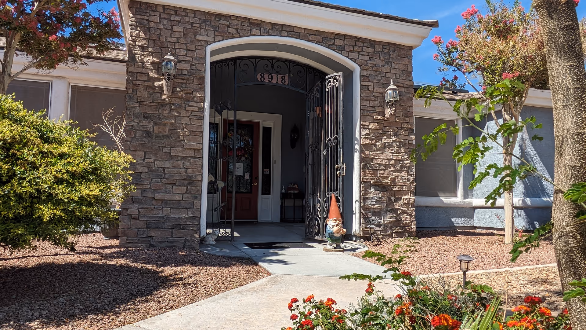 Front entrance of a senior home with a stone facade, an arched doorway with black wrought iron gates, a red door inside, and a garden gnome statue on the pathway. Surrounding the entrance are bushes, small trees, and flowering plants under a clear blue sky.