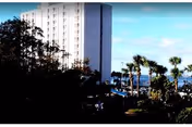 High-rise white residential building beside palm trees overlooking water under a blue sky.