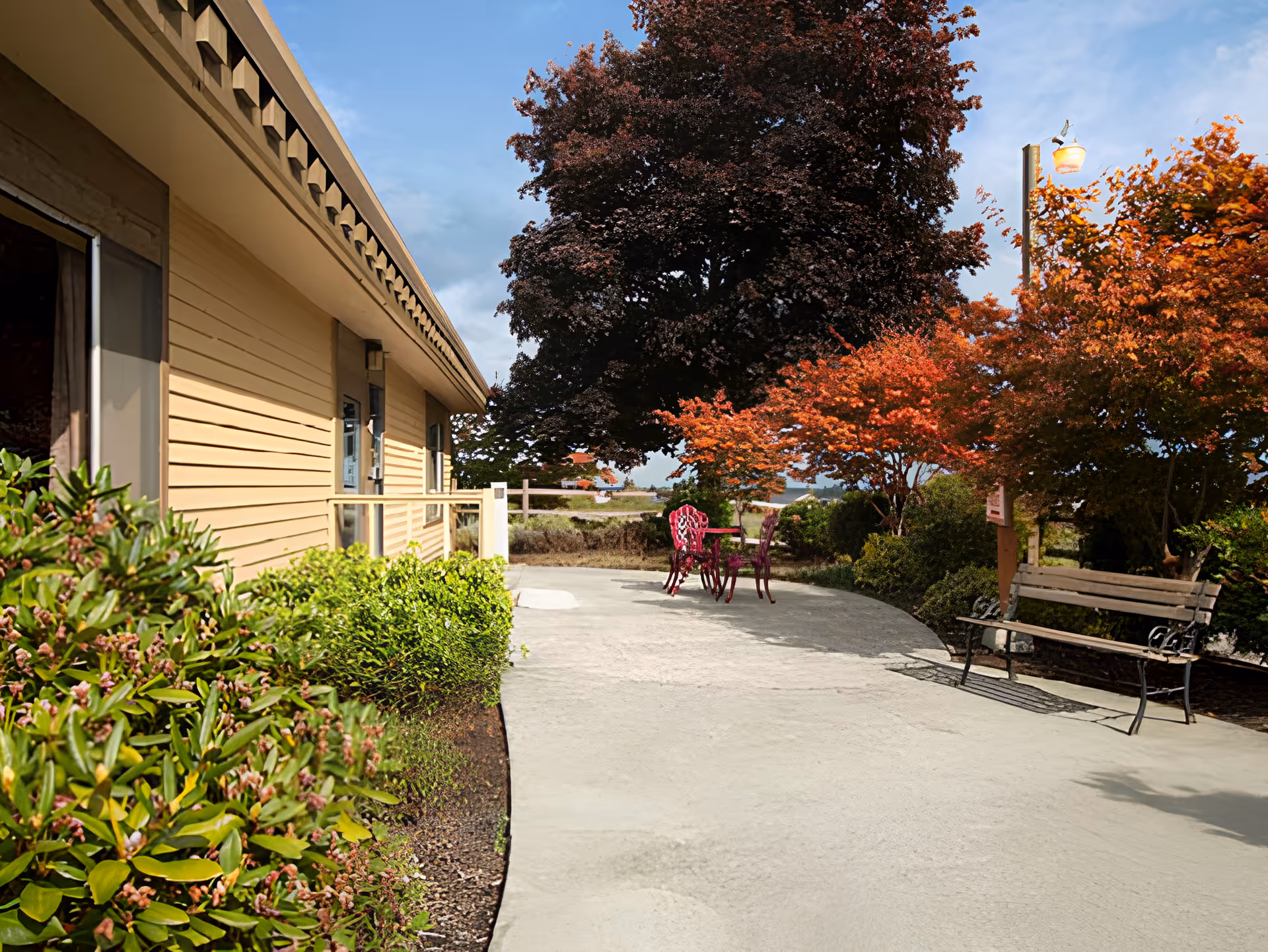 Outdoor patio area next to a beige building with green bushes along the side. The patio has a metal bench on the right and a small table with two chairs further back. Trees with autumn-colored leaves surround the patio under a partly cloudy blue sky.