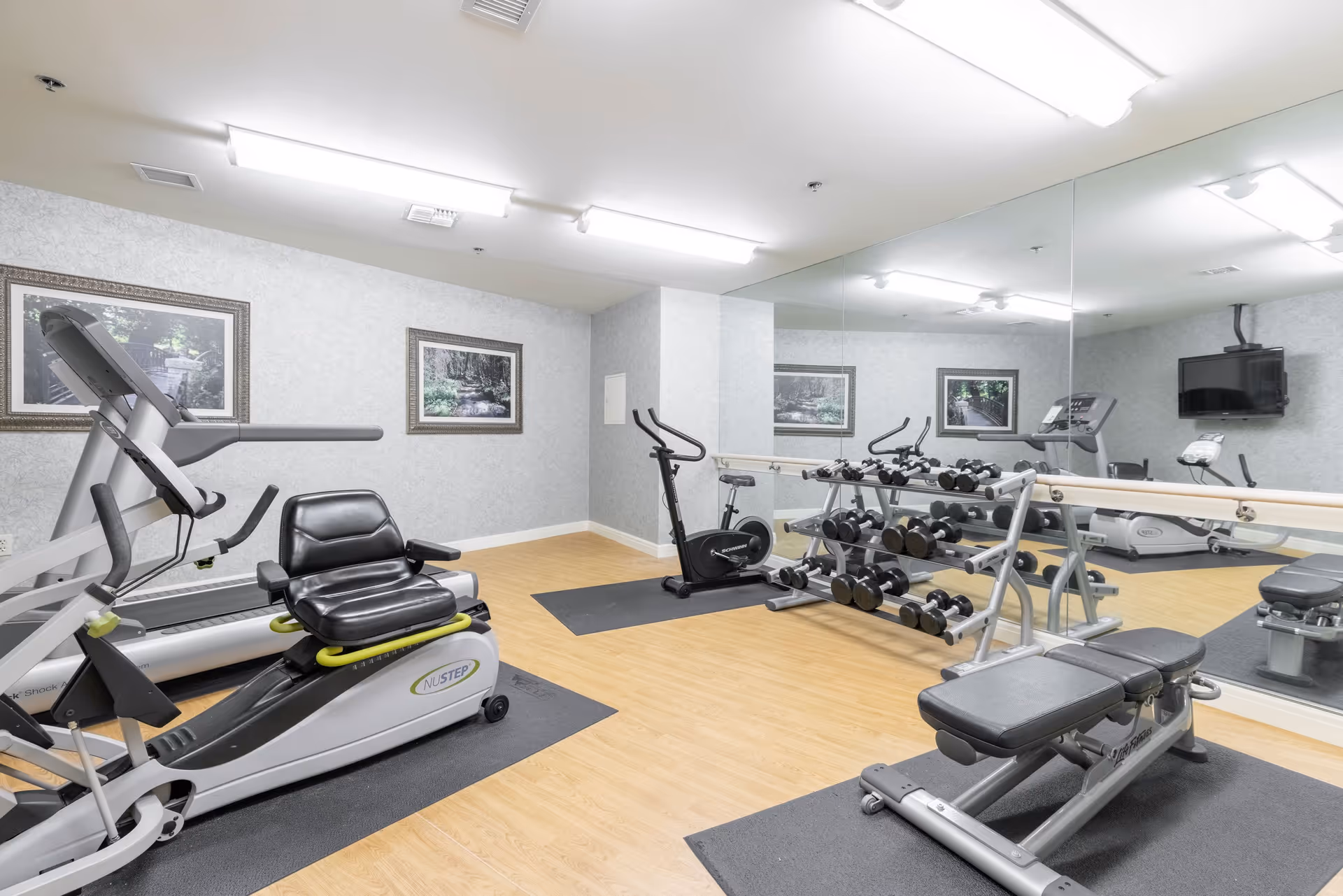 A senior living facility exercise room with various fitness equipment including a recumbent bike, treadmill, stationary bike, free weights on racks, and a workout bench. The room has light-colored walls, wood flooring, large mirrors on one wall, and framed nature pictures hanging on the walls.