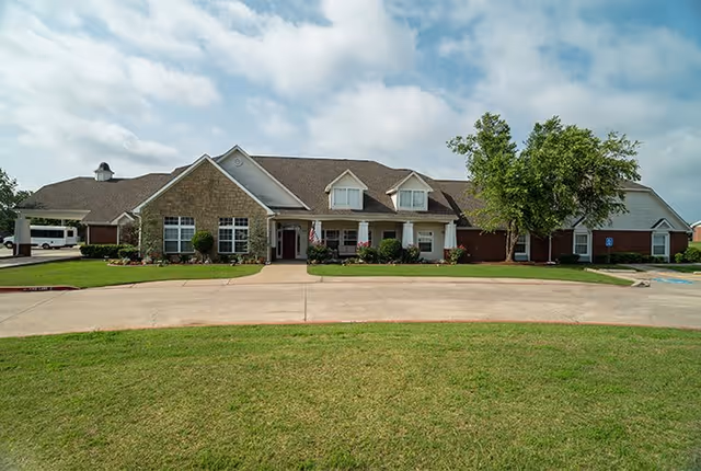 Front exterior view of a single-story senior living facility building with a combination of brick and stone facade, multiple windows, a covered entrance with columns, and a well-maintained lawn and driveway in front under a partly cloudy sky.